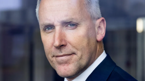 A facial close up of Ciaran Martin looking directly into the lens of a camera. He has got white short hair and is wearing a white shirt underneath a navy suit. The background is blurred.