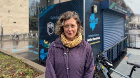 A woman stood in front of an 'Active Travel Hub' outside Ulster University. She is wearing a purple coat and a yellow scarf and has clipped back shoulder length curly brown hair.
