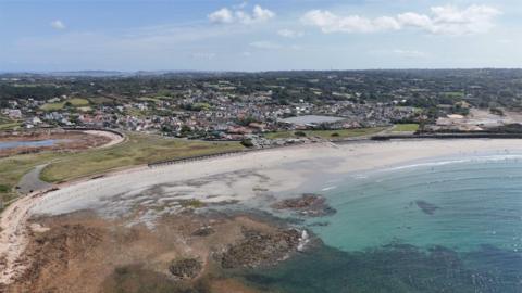 View of Vazon Beach in Guernsey. The water is clear and light blue and there is sand surrounding the sea. Buildings, roads and grass can also be seen in the background.