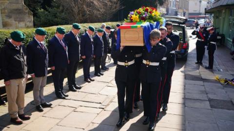 Royal Marine servicemen wearing dark suits with white belts, carrying the wooden coffin of Jim Wren. The coffin is covered with a flag and a large bouquet of spring flowers. Beside the paved walkway stands a long row of former and currently serving marines, wearing green caps and standing with their heads bowed as the coffin passes.