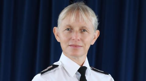 Amanda Pearson poses in front of a blue curtain. She wears a white shirt with epaulettes and a black tie. Her short blonde hair has a fringe.