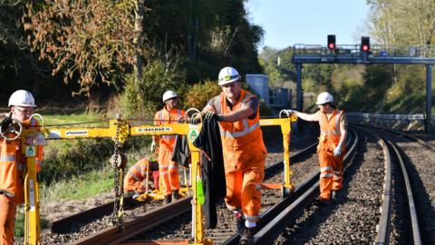 Men in orange hi-vis suits walk down train tracks with a yellow piece of machinery