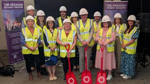 A group of people wearing smart work clothing, with high visibility vests and white hard hats. The three people at the front are holding red shovels. They're all smiling at the camera with banners either side of them about the new Studio Theatre at the Hexagon.