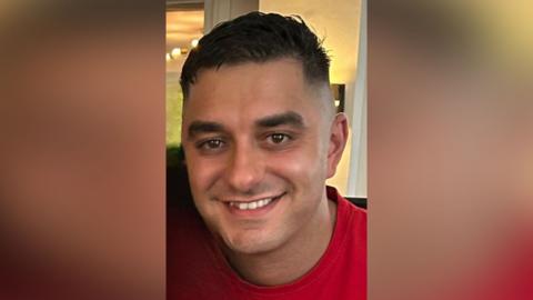 Jordan Hogg smiles at the camera. He is clean shaven with large brown eyes and short dark hair.