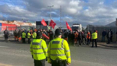 Two police officers face a picket line outside the entrance to a bin depot. People, some who are holding a red, Unite the Union, flag are lined up in front of steel gates. The sky is blue and cloudy in the background.