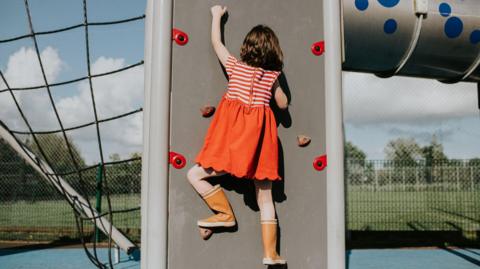 A little girl scales a climbing wall in a play park on a sunny day.