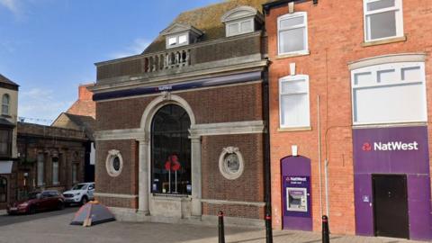 The closed NatWest bank in Cleethorpes. A grand-looking dark-brick building has decorative stonework and a tall arched window, with smaller circular windows to either side. To the right is a less distinctive, red-brick, terraced building, featuring a large NatWest sign on purple boarding, and a cash machine.