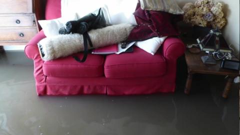 A red sofa with belongings piled on top of it. It is surrounded by floodwater which looks grey and has flecks of white in it.