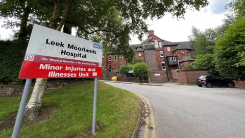 A red and white sign under a tree on the left says "Leek Moorlands Hospital - Minor Injuries and Illnesses Unit". A brick building with walls around it can be seen in the background.