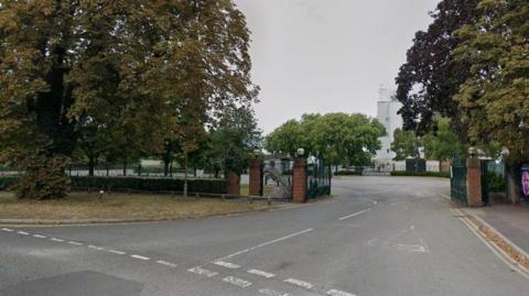 The entrance to Kempton Park racecourse seen in a Google Streetview image. There are large metal gates which are open and one of the stands can just be seen behind some trees.