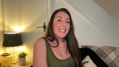 Amie Wood sits and smiles at the camera. She has long brown hair and wears a dark green tank top. She is sat in a living room, with a door, coffee table, lamp, and plant pot behind her to her right.