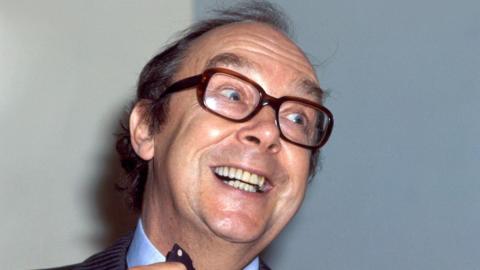 Eric Morecambe looks to the side of the camera. He is smiling and straightening his tie. He wears brown, thick-framed glasses. He has short dark hair.