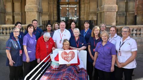 A group of nurses and hospice staff in uniform gather around a woman sitting in a hospital bed. The woman has her legs covered by a chequered blanket and is holding sign with a big red heart and the words 'Thank you!' written on the front.