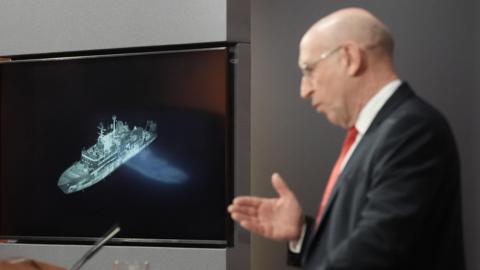 John Healey at a lectern with a picture of the Yantar behind