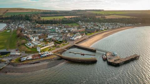 An aerial view of the village showing its harbour, with boats hauled up on a nearby shore, and white-walled properties. There are trees and fields behind the village.