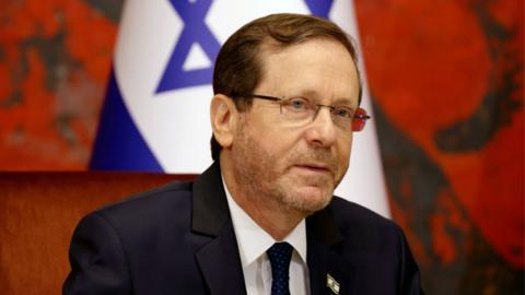 A man with short brown hair and glasses, in a navy suit, pictured in front of a white and blue Israeli flag with a star