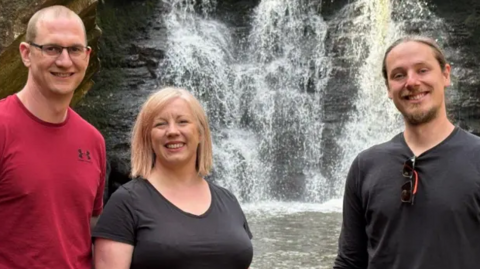 Two men and one woman - they look youngish in their 30s- standing in front of a waterfall.