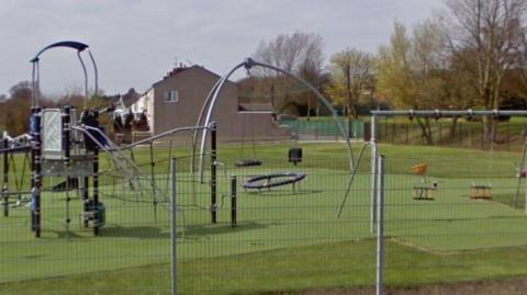 A general view of a play area with swings, a slide and other play equipment. The play park is near some houses, which are visible in the background.