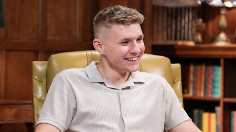 A man sitting in a yellow leather chair in an old looking room with dark wood walls and bookshelves. He is looking off-camera and smiling. He has short brown hair and a nose ring, and wears a beige short-sleeved shirt.