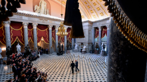 A wide view shows the King being led through the hall of the Capitol with reporters to the side.