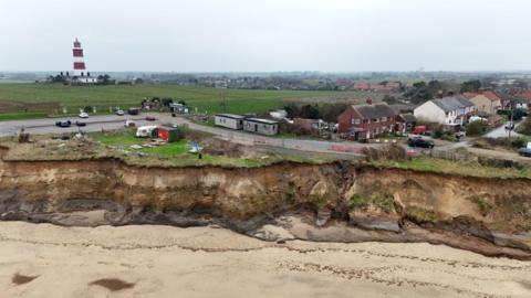 The cliff edge at Happisburgh. The cliffs are very sandy and there is a beach below. Houses and cars are very close to the cliff edge. There is a red and white striped lighthouse in the distance.