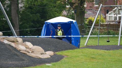 A police officer stands outside a blue tent which is in a playground, behind blue tape.
