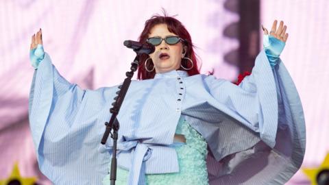 CMAT performs on Radio 1's Big Weekend stage against a pink backdrop with yellow stars. She wears a light blue floral two-piece outfit with a blue striped cape and has shoulder length red hair. She wears silver hoops and sunglasses and holds up her hands to the crowd