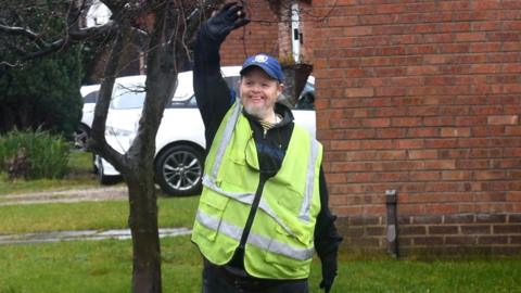 Randal wearing a high visibility yellow jacket with a dark top underneath and a blue baseball cap with a white log on it. He is holding his right arm up and waving
