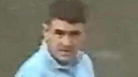 CCTV footage still shot of a man with brown hair. He is wearing a light blue half zip. 