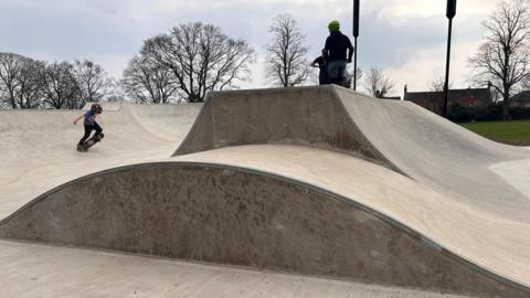 Three people on skateboards and helmets skating on a curved surface in a park.