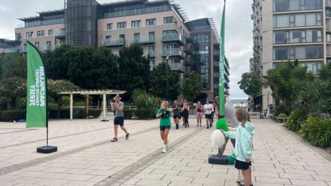 Runners participating in an event on a paved urban pathway, flanked by modern buildings and greenery. Two green 'Macmillan Cancer Support' flags are visible on the left. A child in a light blue jacket stands on the right, cheering with white pom-poms. The sky is overcast