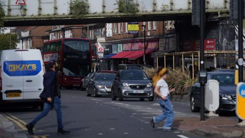 Two people cross a road in Golders Green, with cars, a bus and shops in the background