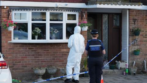 A police community support officer and a forensic examiner in a white suit are outside a modern red brick property, which has been sealed off with blue and white police incident tape. 