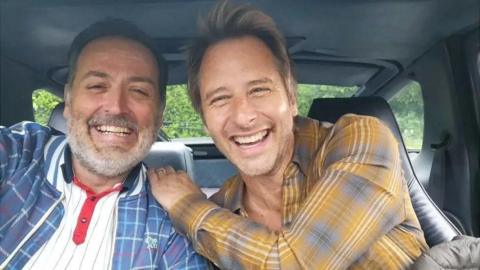 Vic Irvine and Chesney Hawkes are sat in the front seats of a car smiling at the camera. Mr Irvine sits to the left and has black hair flecked with greys and a grey beard. He wears a white polo top with black stripes and red detailing on the button strip and a blue tartan print bomber jacket over the top. Chesney Hawkes has his left hand on Mr Irvine's shoulder, he has blonde short hair and wears a mustard yellow tartan print shirt.