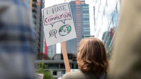 Woman protests outside Amazon office with a sign which reads 'I hate commuting'.