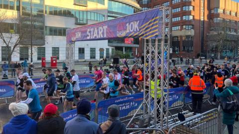 Runners are seen starting a 10K race under a large banner that reads “START”. The scene includes a crowd of participants in athletic gear moving forward, while event staff in bright orange and yellow vests manage the area. Metal barriers line the route, and spectators watch from the sides.