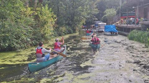 People in several turquoise canoes paddle along a river past moored boats and a quayside area, and trees on the opposite bank.