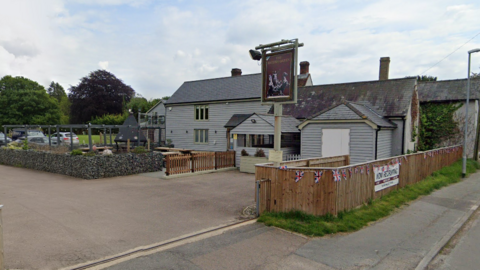 A car park is on the left of the picture with 4 cars. There is outdoor seating in front of the pub, with a wooden fence cornering it off. The pub is grey and has wooden cladding on the outside. It also has a sign outside that says British Queen on it. 