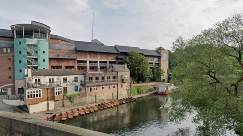 Prince Bishops Place's multi-storey car park is mostly made of brown bricks and overlooks the River Wear. One of its walls is bright blue. There are about 20 rowing boats on the side of the river. 
