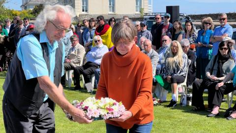 Two people who a wreath as they are about to lay it down as a large crowd watches them from behind