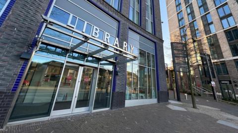 A tall, brick-built building with metal letters spelling out the word 'library' above a set of glass automatic doors. The building is situated next to another tall building and there are trees and street lights visible nearby.