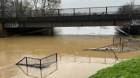 A river floods its banks with brown water. The water level is almost reaching a bridge over the river, and bankside fencing is submerged.