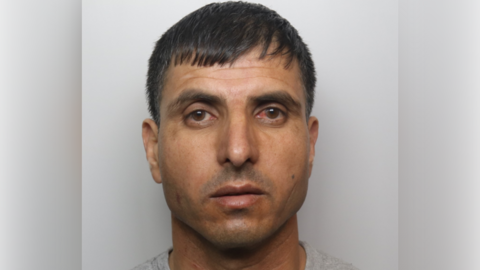 A police mugshot of a man with short black hair and wearing a grey top.