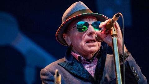 Van Morrison on stage. He is wearing a hat, a jacket and a patterned pink shirt. He is also wearing sunglasses and holding a microphone. 