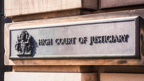 A sign on a sandstone block exterior wall. The sign has a royal crest and reads: "High Court of Justiciary".