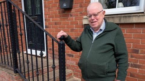 An elderly man in thinning white hair stands outside a red brick house holding on to a black railing