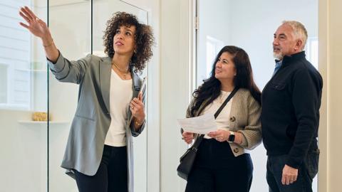 A stock photo showing an estate agent gesturing as she shows a couple a bathroom as part of a house viewing