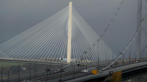 The Queensferry Crossing suspension bridge stands behind the Forth Road Bridge with a grey sky in the background. 