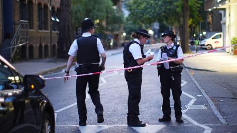 Three uniformed police officers - one male and two female - stand in a road behind police tape.