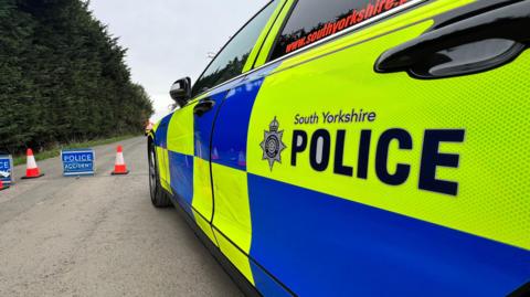 A police car with fluorescent yellow and bright blue branding is parked by a blue police sign and road cones.